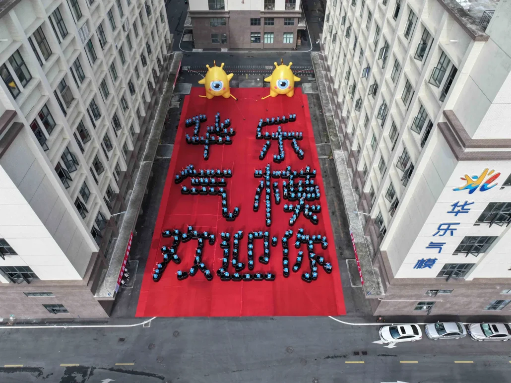 Drone view of a courtyard between buildings displaying cars arranged in patterns on a large red carpet with inflatable figures.