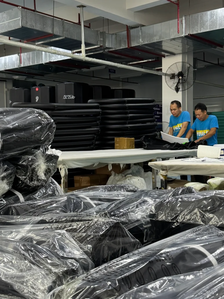 Factory setting with two workers at a table reviewing items, surrounded by packaged black products and industrial equipment.
