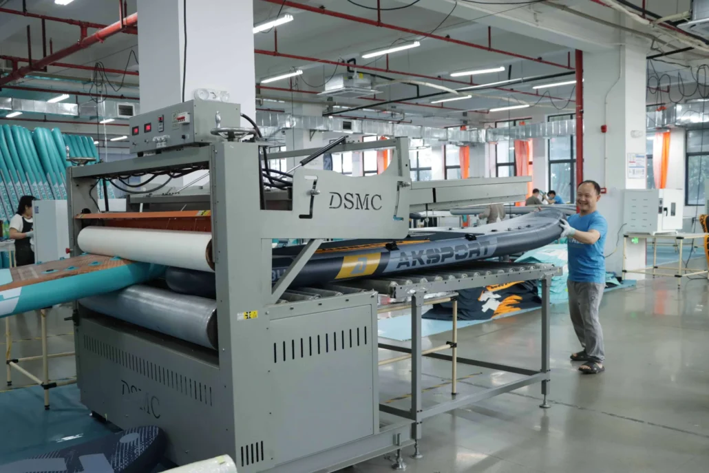 Factory machine labeled DSMC processing inflatable paddleboards, with a worker handling a board marked 'AXSPORT'.