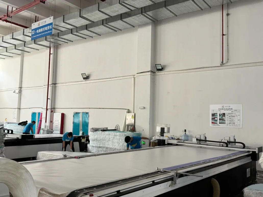 Workers in a factory operating large CNC cutting machines, with sheets and rolls of material visible. Industrial setting with signage.