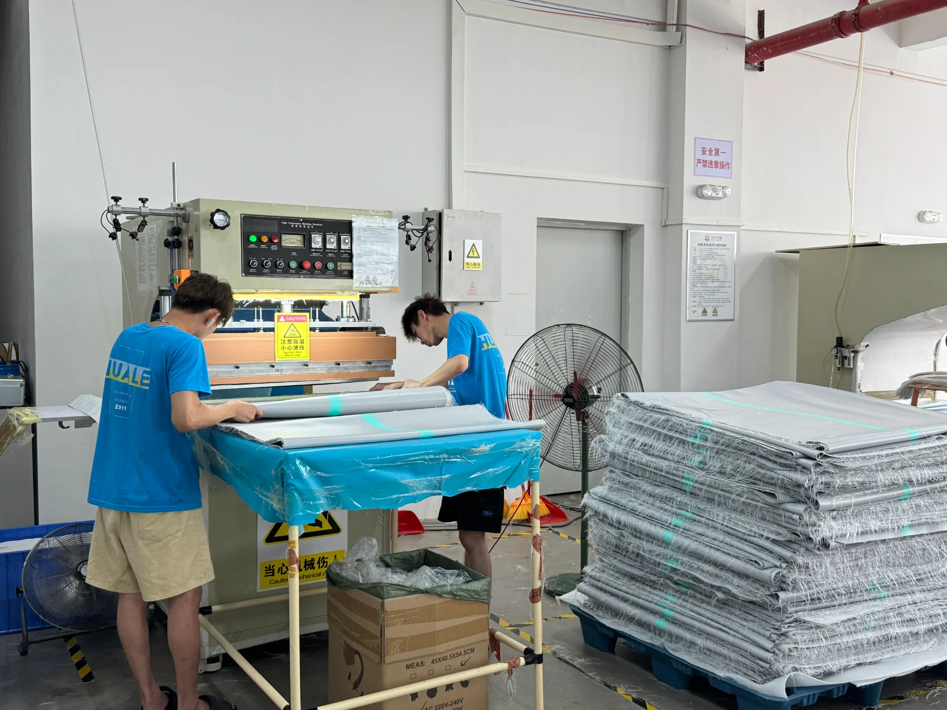 Two workers in blue shirts operate a machine in a factory, with folded materials stacked beside them.