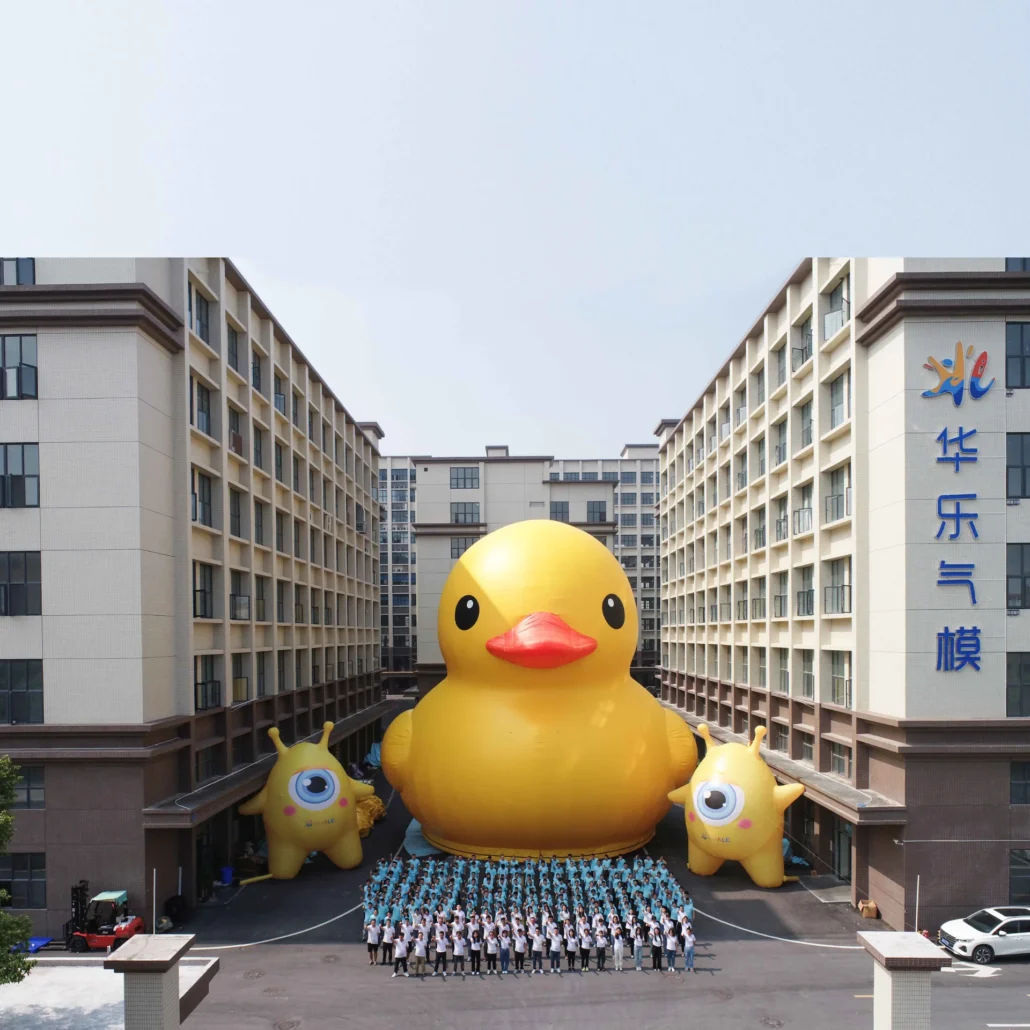 Large inflatable rubber duck surrounded by people and smaller inflatables outside a building with tall walls.