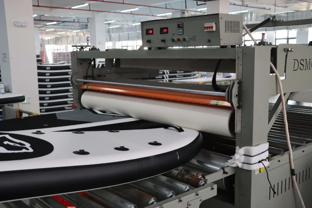 Close-up of machinery used to produce inflatable paddleboards in a factory setting, showcasing industrial equipment and paddleboards.