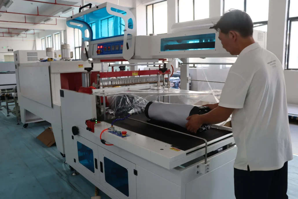 Worker operating a large industrial packaging machine in a factory, handling a rolled product for wrapping.