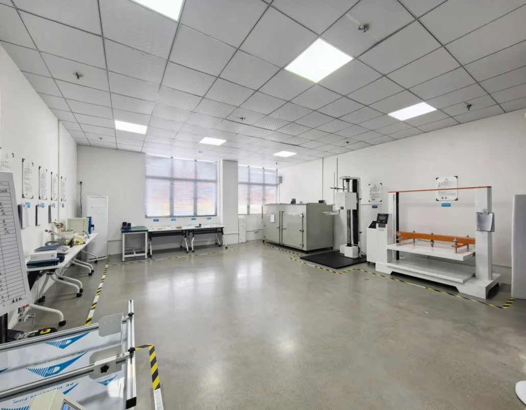 Spacious industrial testing lab with various equipment, including testing machines and workstations, under a grid ceiling and large windows.