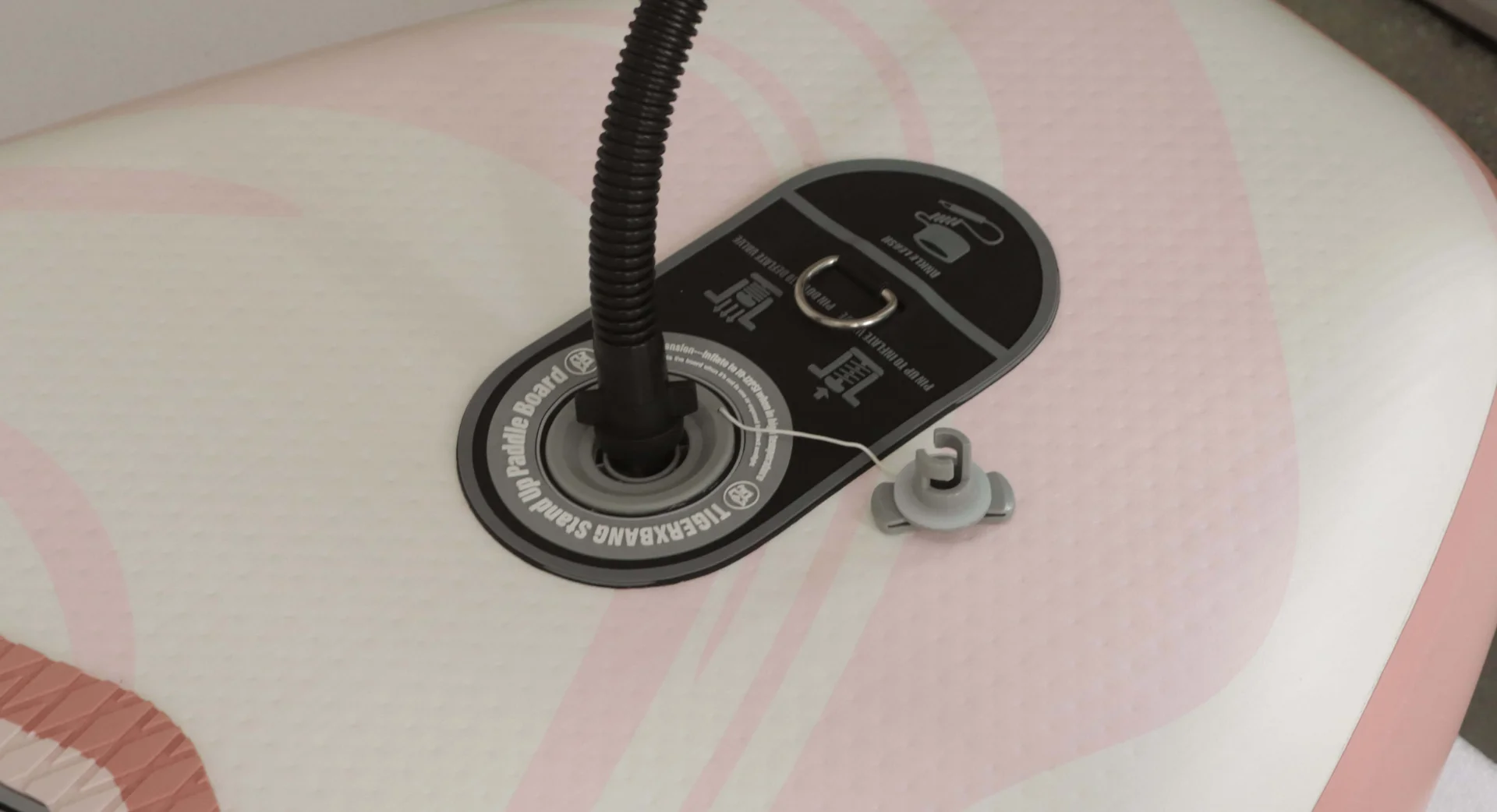 Close-up of an inflatable paddle board valve, a pump hose attached, and a cap on a pink and white surface.