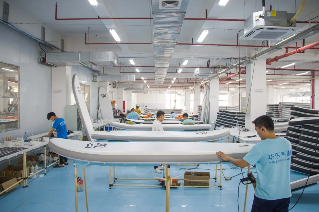 Workers in a factory assembling inflatable paddleboards, surrounded by paddles and equipment under bright ceiling lights.