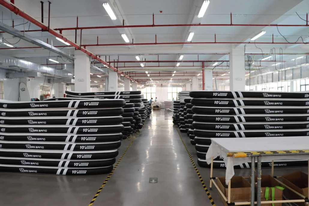 Rows of black inflatable paddleboards stacked in a warehouse setting, under bright ceiling lights.