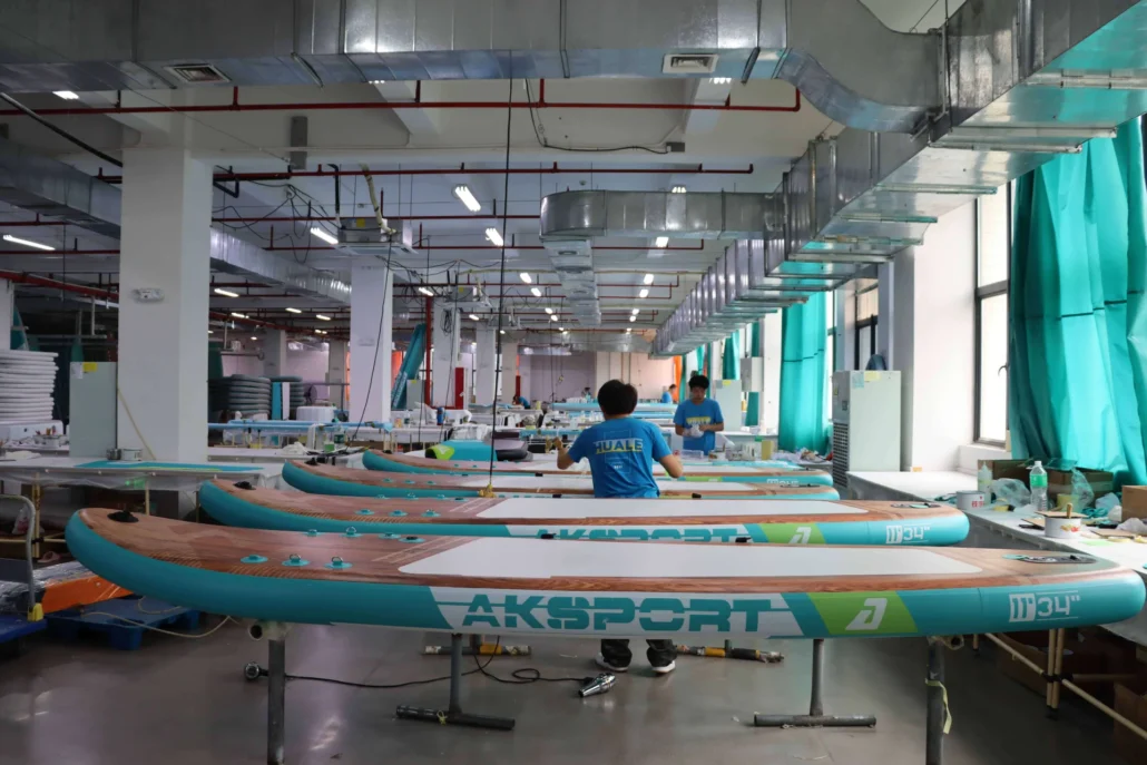 Workers assembling inflatable paddleboards in a factory with multiple boards and industrial equipment visible. Bright lighting from overhead fixtures.