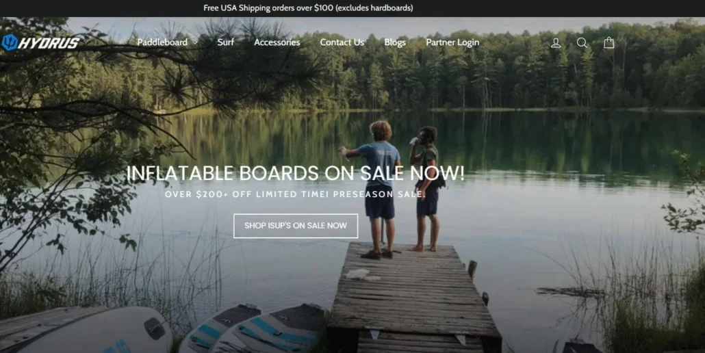Two people standing on a dock by a lake surrounded by trees, with inflatable paddleboards nearby and promotional text about a sale.