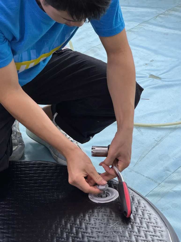 Individual in a blue shirt using a tool to repair an inflatable by tightening a valve on a textured surface.