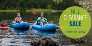 Two people kayaking on a lake surrounded by trees, with a sign promoting a fall clearance sale and a call to action to explore deals.