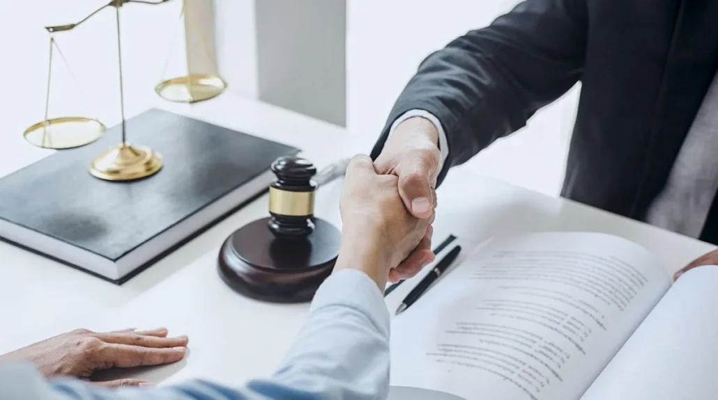 Two people shaking hands over a legal agreement on a desk, with a gavel, scale, and pen visible, symbolizing a business or legal setting.