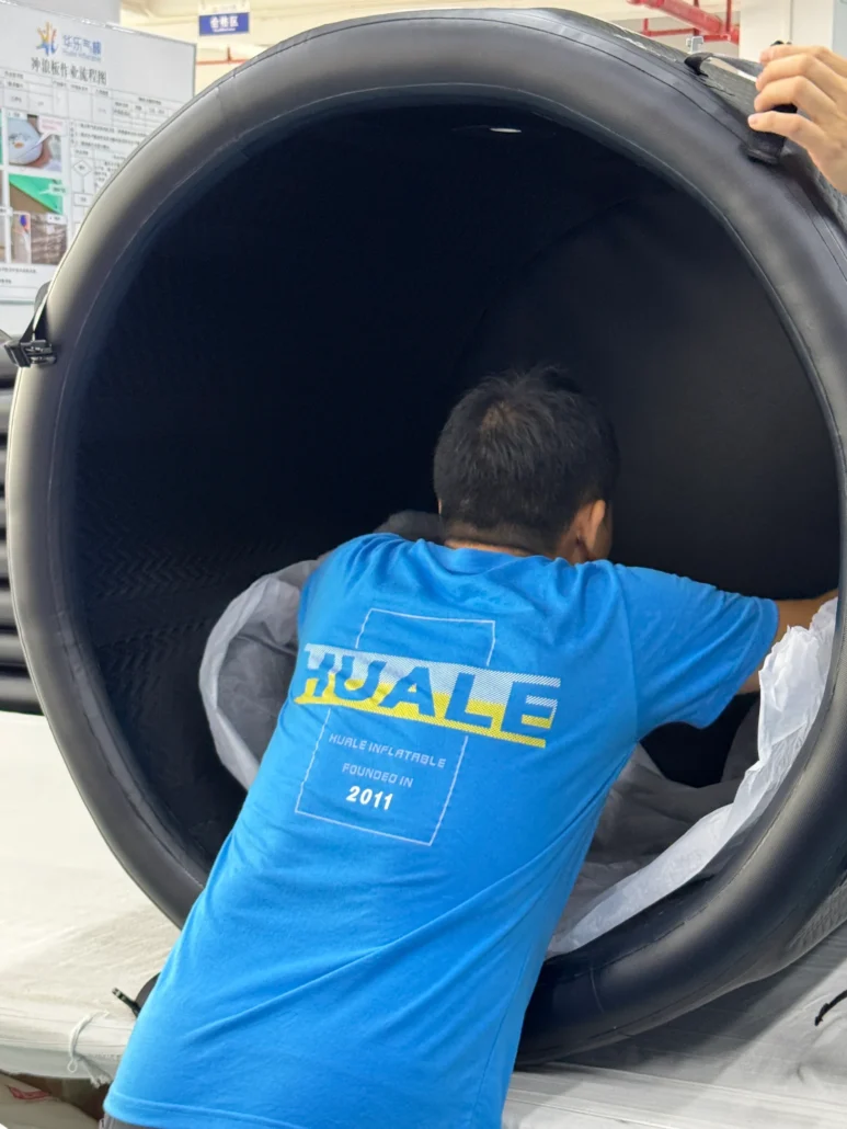 A man in a blue Huale branded shirt inspecting the interior of a large black inflatable tube in an indoor setting.
