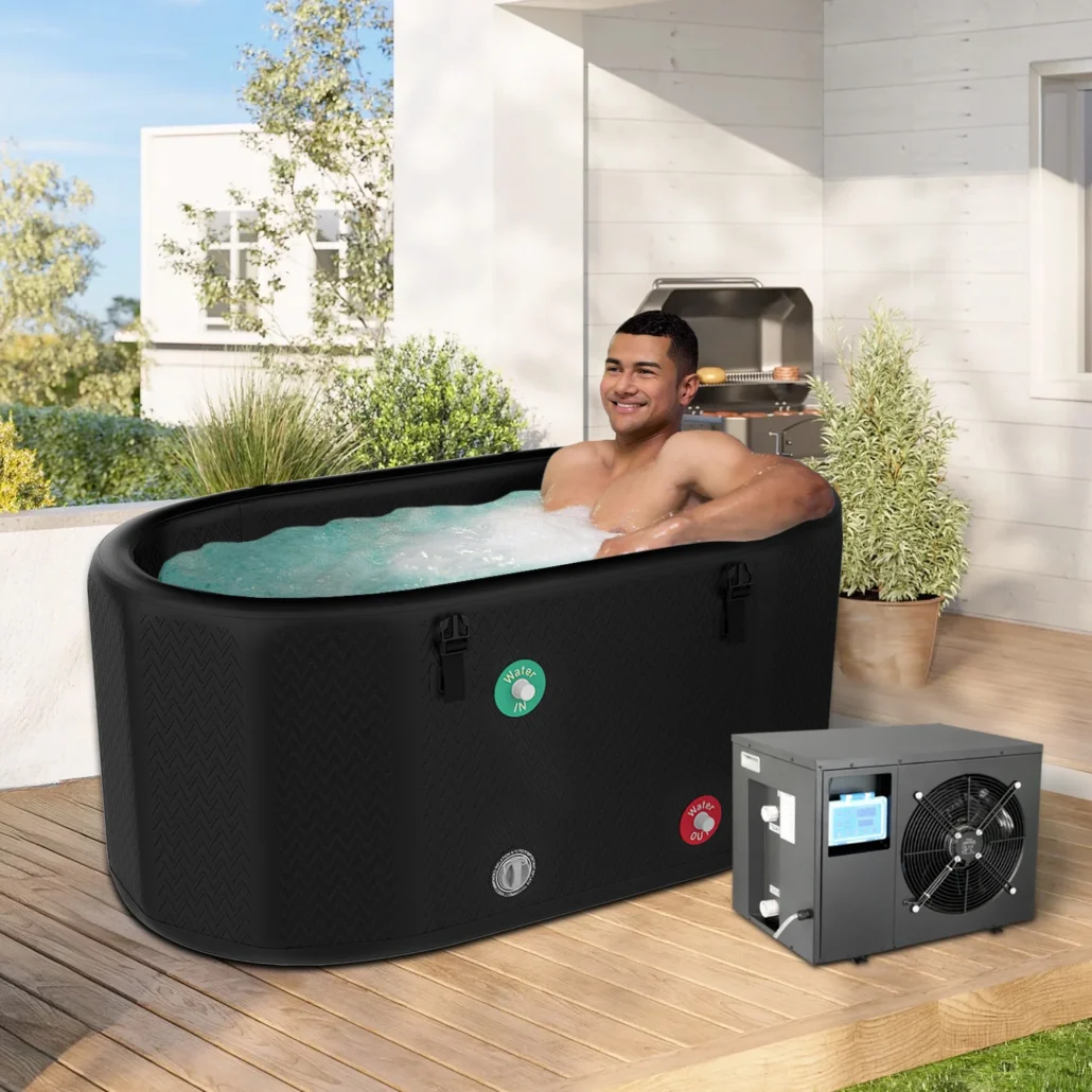 A man lounging in a black portable hot tub filled with water on a wooden patio, next to a heater unit and outdoor barbecue grill.