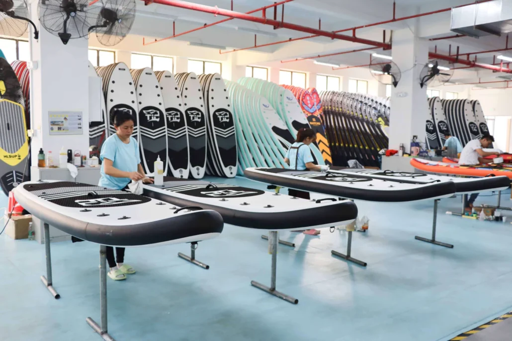 Workers in a factory assembling and fine-tuning paddleboards, with stacks of boards in the background.