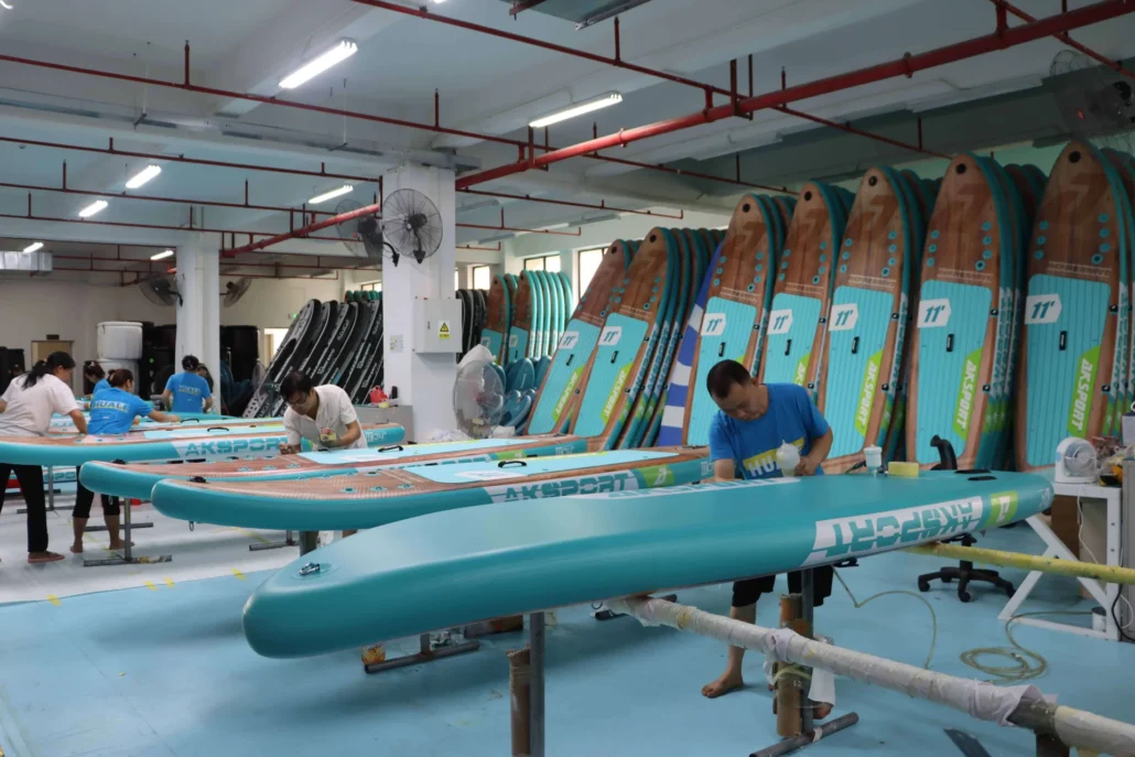 Workers manufacturing paddleboards in a factory with multiple boards in production stages, surrounded by equipment.