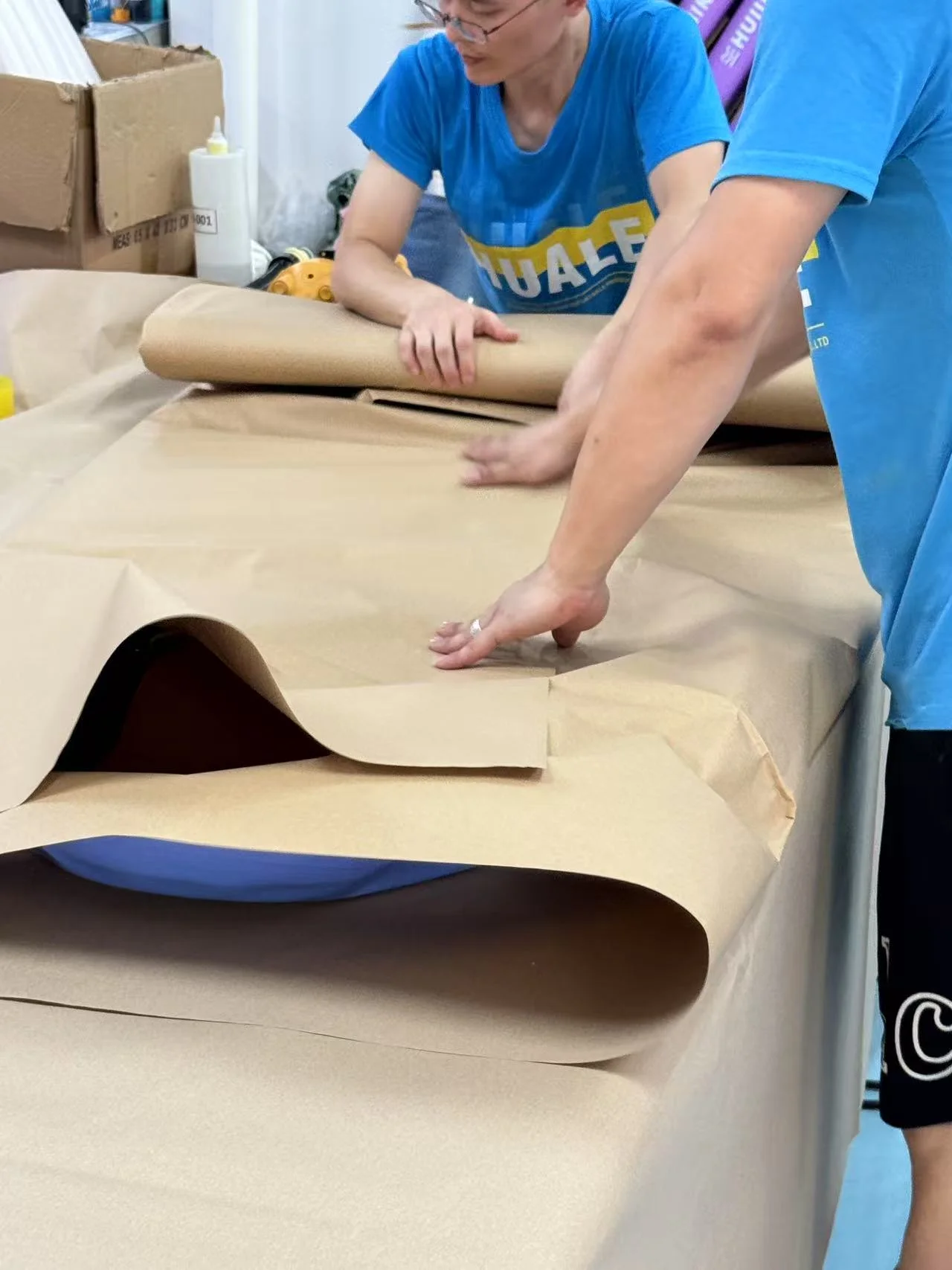 Two workers wrapping a large object in brown paper on a table, with tools and materials around them.
