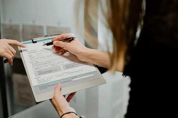Person holding a clipboard while filling out a form with a pen, receiving guidance from another person pointing at the document.