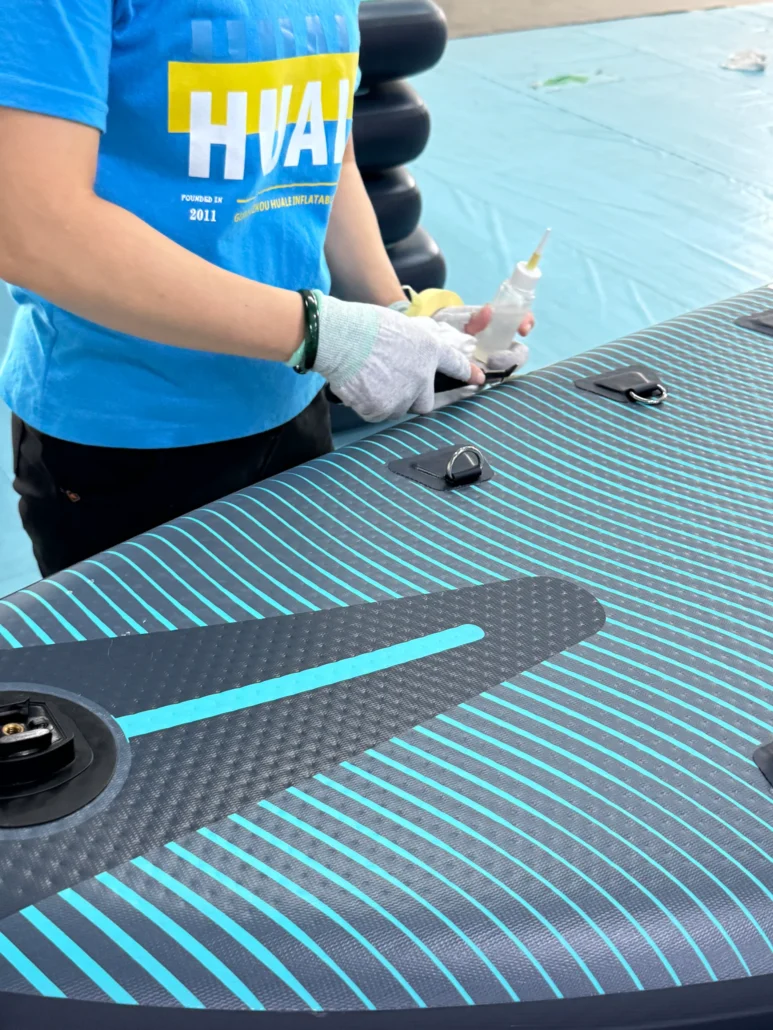 Individual in blue shirt and gloves applying glue to an inflatable paddleboard with striped pattern on a light blue surface.