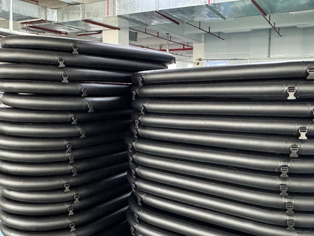 A large stack of black PVC pipes piled orderly in an industrial space with exposed ducts and pipes visible in the background.