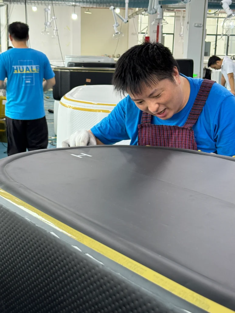 Factory worker wearing a blue shirt assembles a bathtub in a manufacturing facility, with tools and equipment in the background.