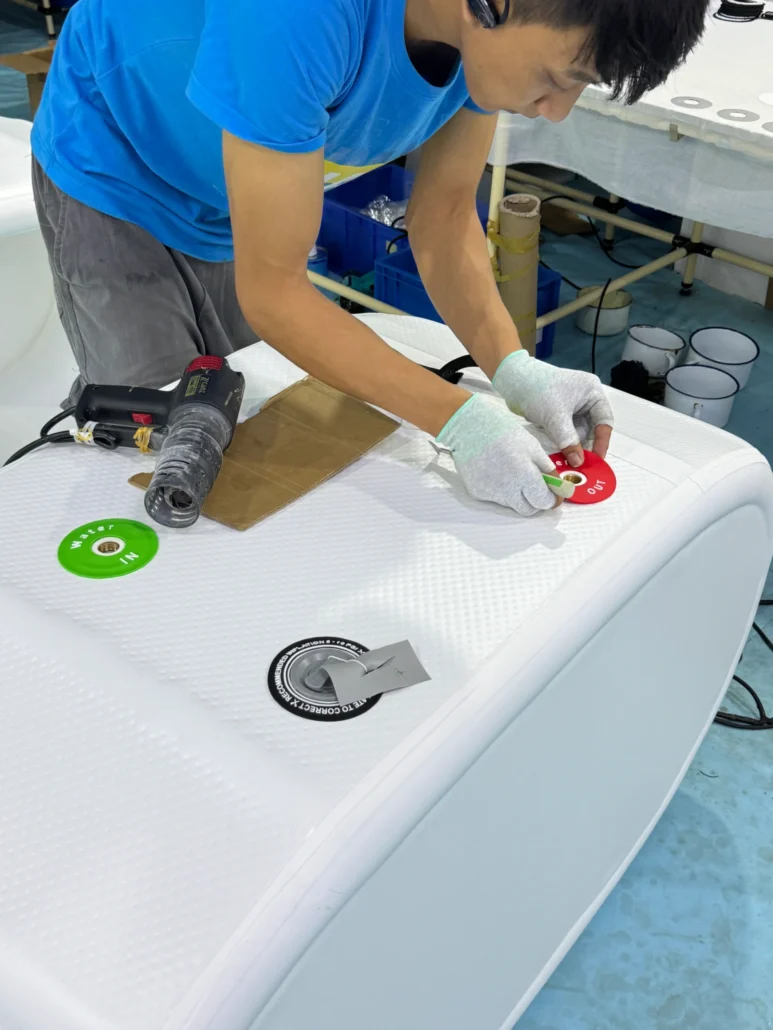 Person in blue shirt and gloves attaching components to a white inflatable product using tools on a workbench.