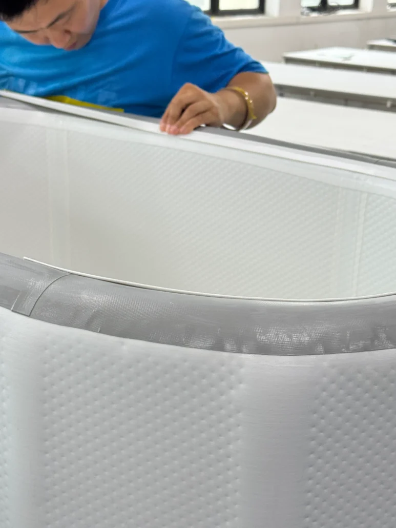 A worker in a blue shirt carefully inspecting the edge of a white quilted mattress in a factory setting.