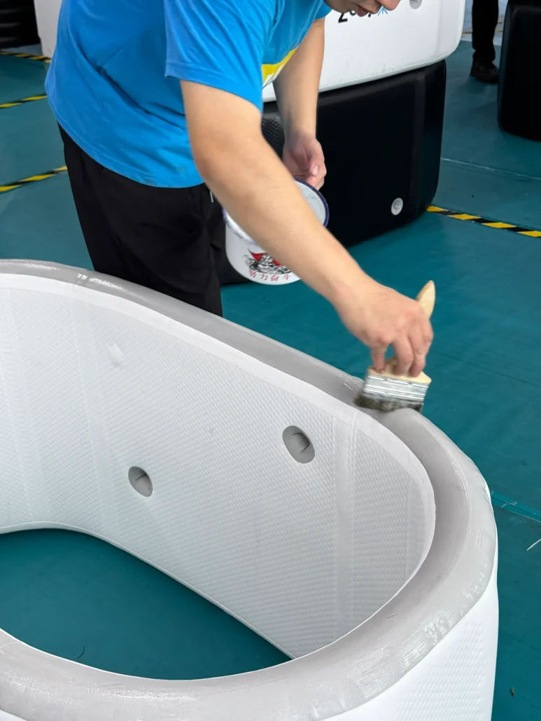 A person in a blue shirt applies paint to the edge of a white inflatable pool using a brush. The setting is indoors with a teal floor.