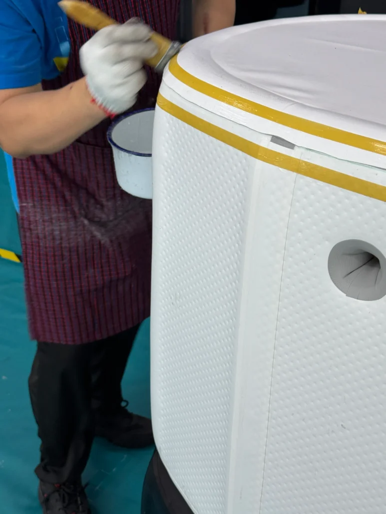 Worker painting a white textured surface using a brush, wearing gloves and apron, with masking tape applied for precision.