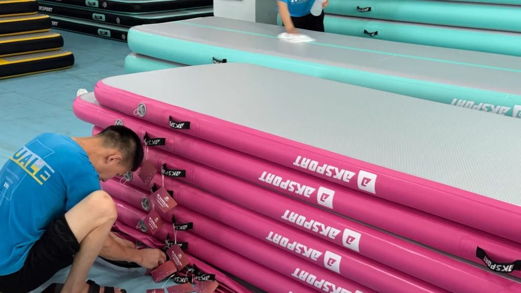 Two workers preparing colorfully stacked airtrack mats, one attaching labels to pink mats and another cleaning teal mats.