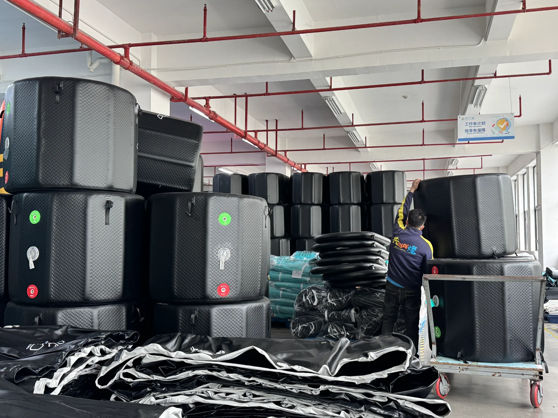 A factory worker organizing large black inflatable products in a storage area. Stacked items, rolls of material, and equipment are present.