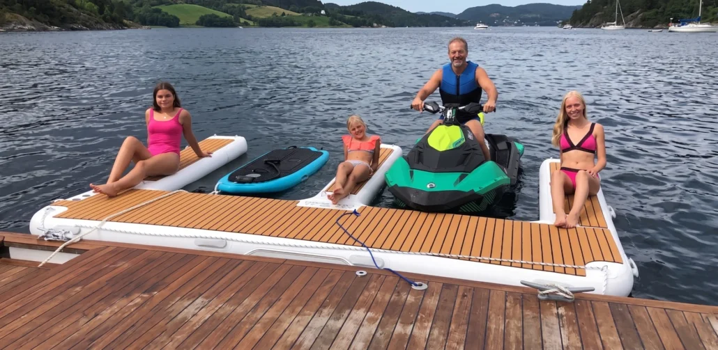 A family relaxing on a wooden floating dock on a calm lake, featuring a green and black jet ski and inflatable platforms.