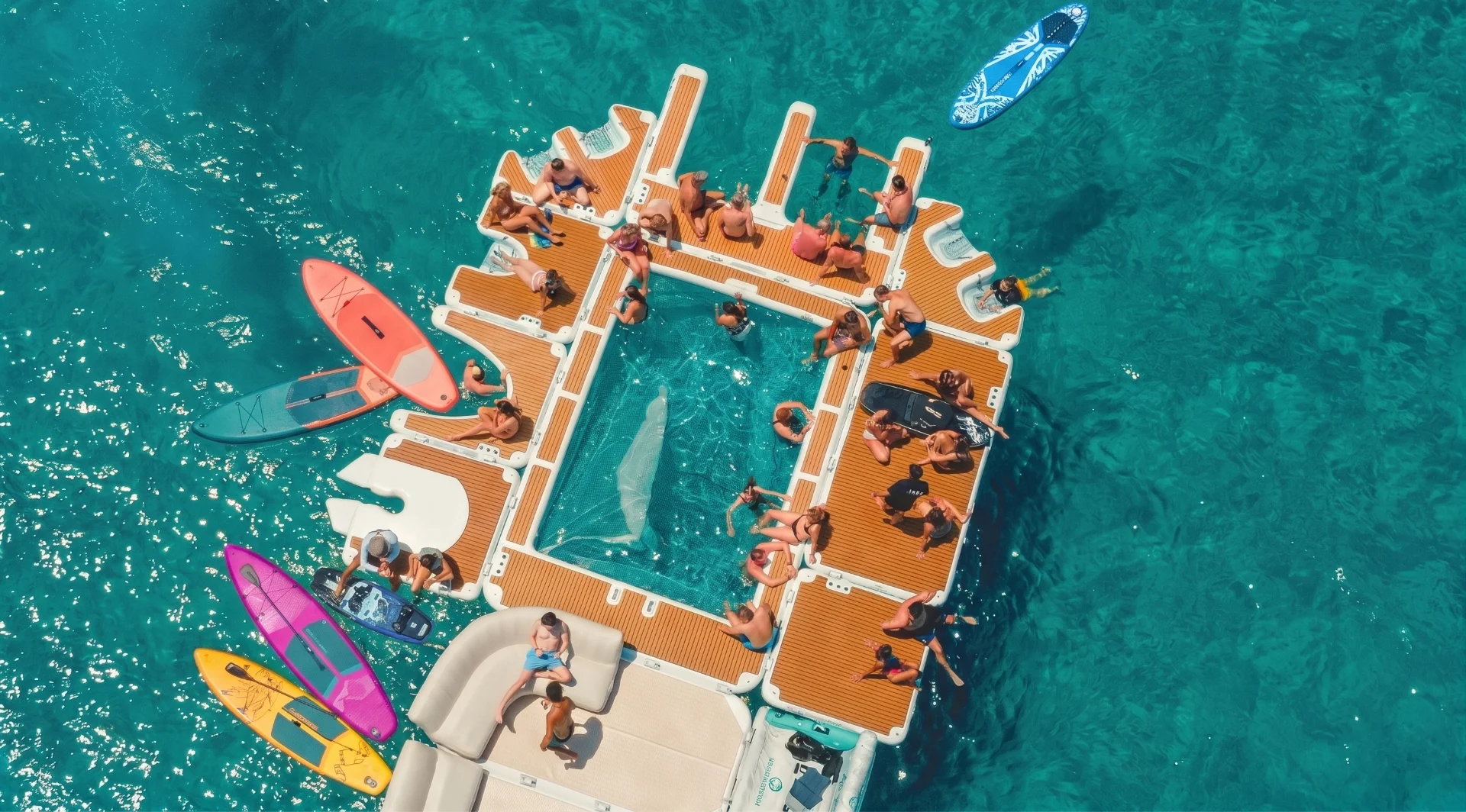 A group of people enjoying a party on a floating deck with wooden panels and a central net enclosure, surrounded by paddleboards in clear turquoise tropical water.