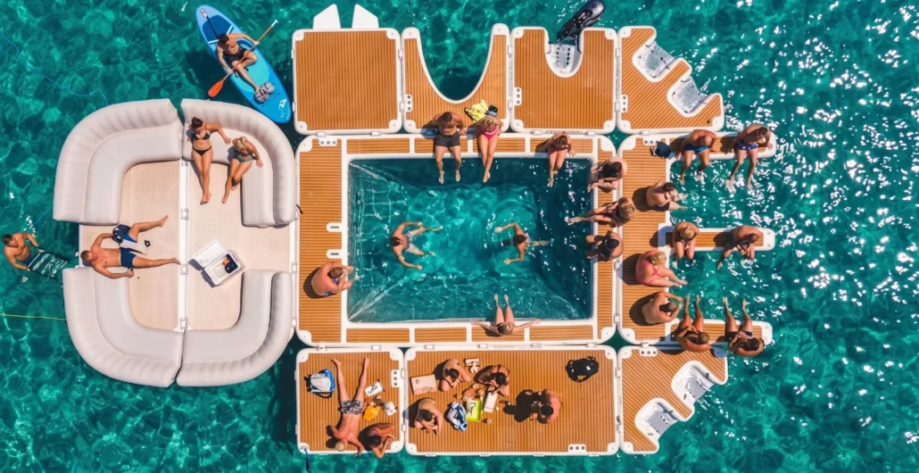 Aerial view of a floating party deck with multiple sections and people lounging or swimming in a calm, crystal-clear body of water.