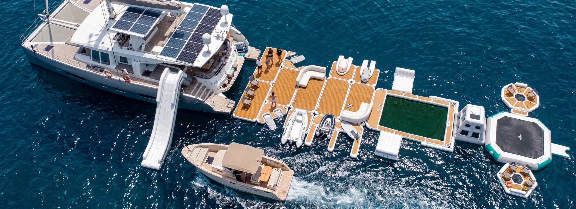 Overhead view of a luxury yacht with extended floating platforms featuring seating areas, boats, a netted swimming area, and other recreational setups in open water.
