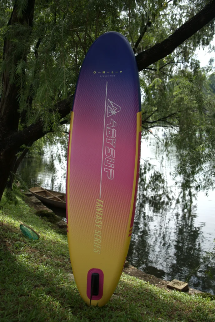 A vibrant pink, yellow, and blue paddleboard labeled 'Fantasy Series' propped against a tree by a calm riverside, with a wooden boat in the background.