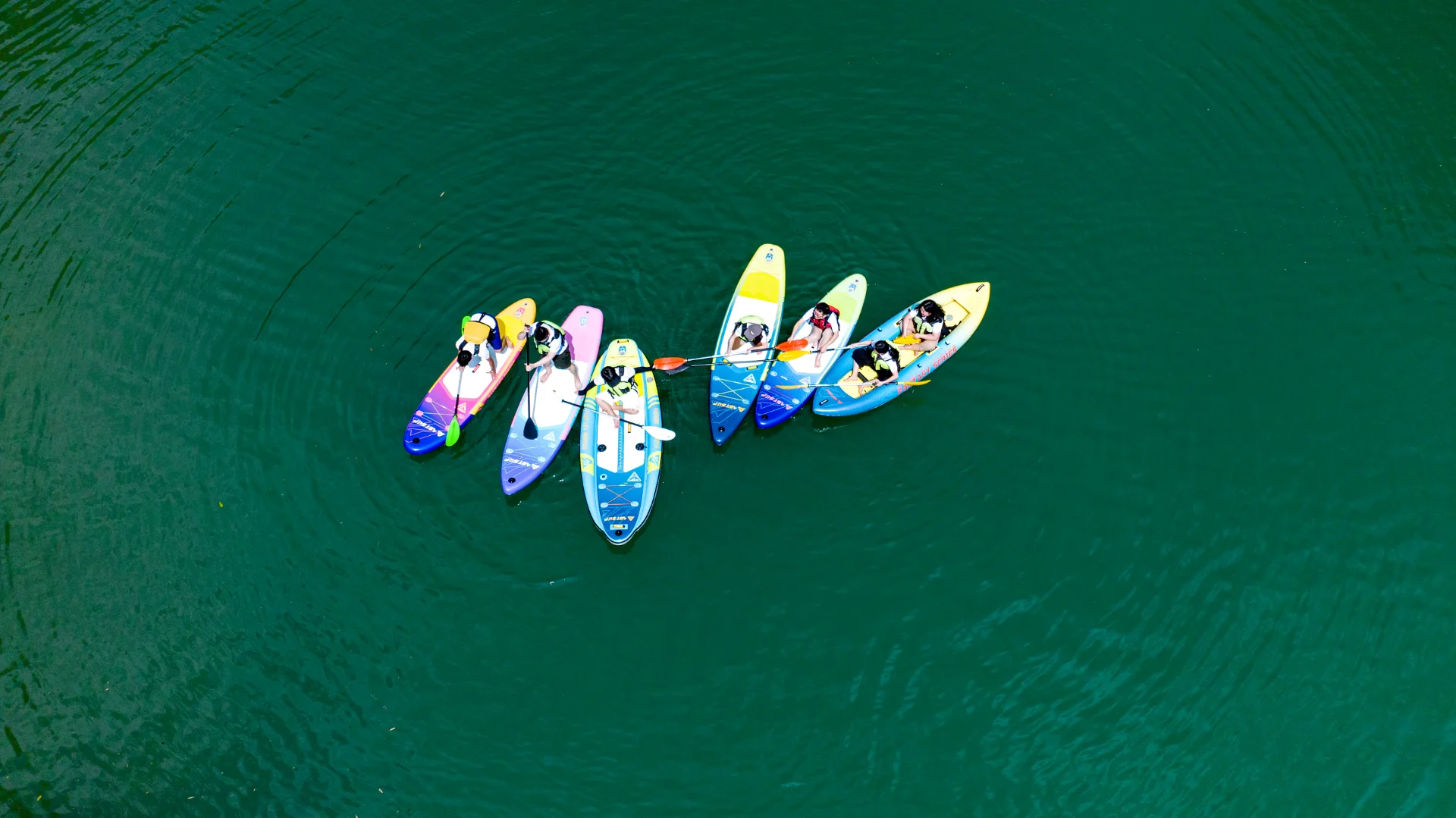 Aerial view of a group of people paddleboarding on calm green water, their boards of various colors forming a semicircle.