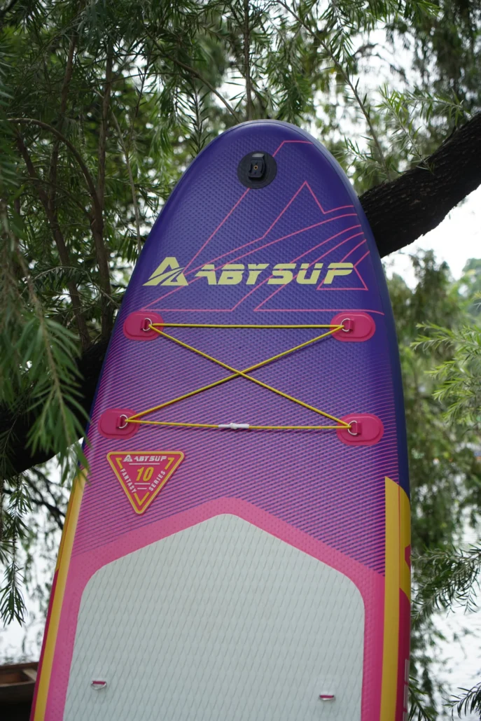 Close-up of a purple stand-up paddleboard with vibrant designs, bungee cord storage, and visible logo, hanging in a tree.