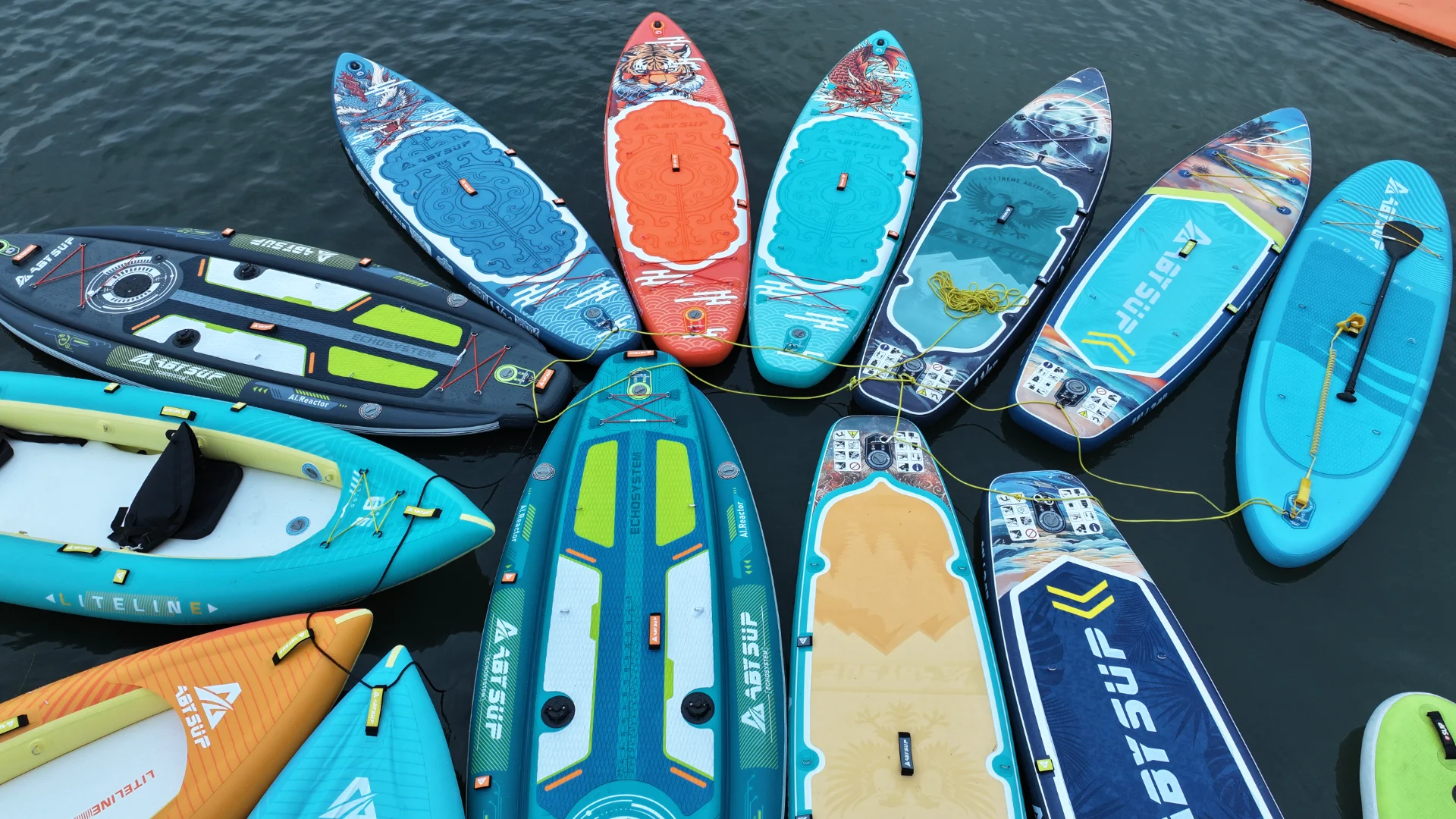 A close-up view of various colorful stand-up paddleboards and inflatable kayaks tethered together on a calm water surface.