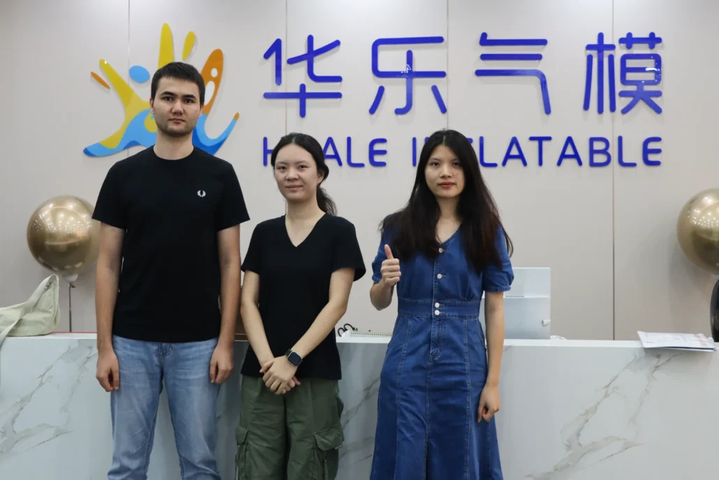 Three individuals standing in front of a reception desk with the Huale Inflatable logo on the wall behind them, one giving a thumbs-up gesture.