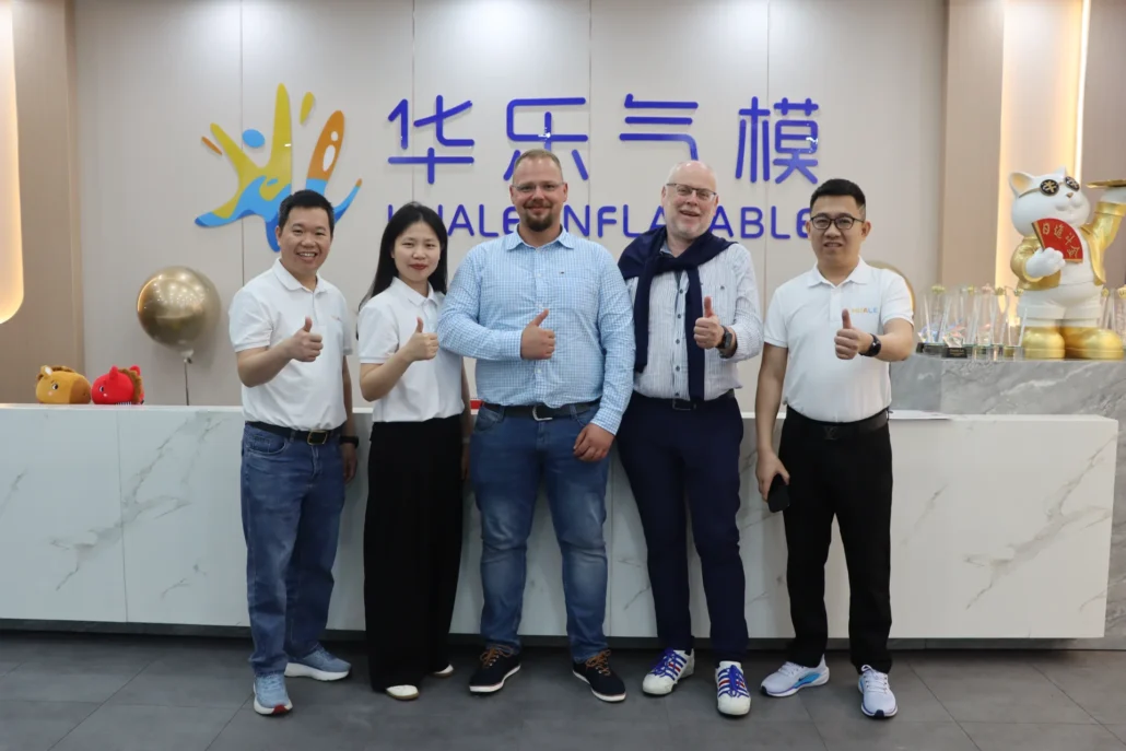 A group of five individuals posing together in front of a company logo, smiling and giving thumbs up, with trophies and decor in the background.