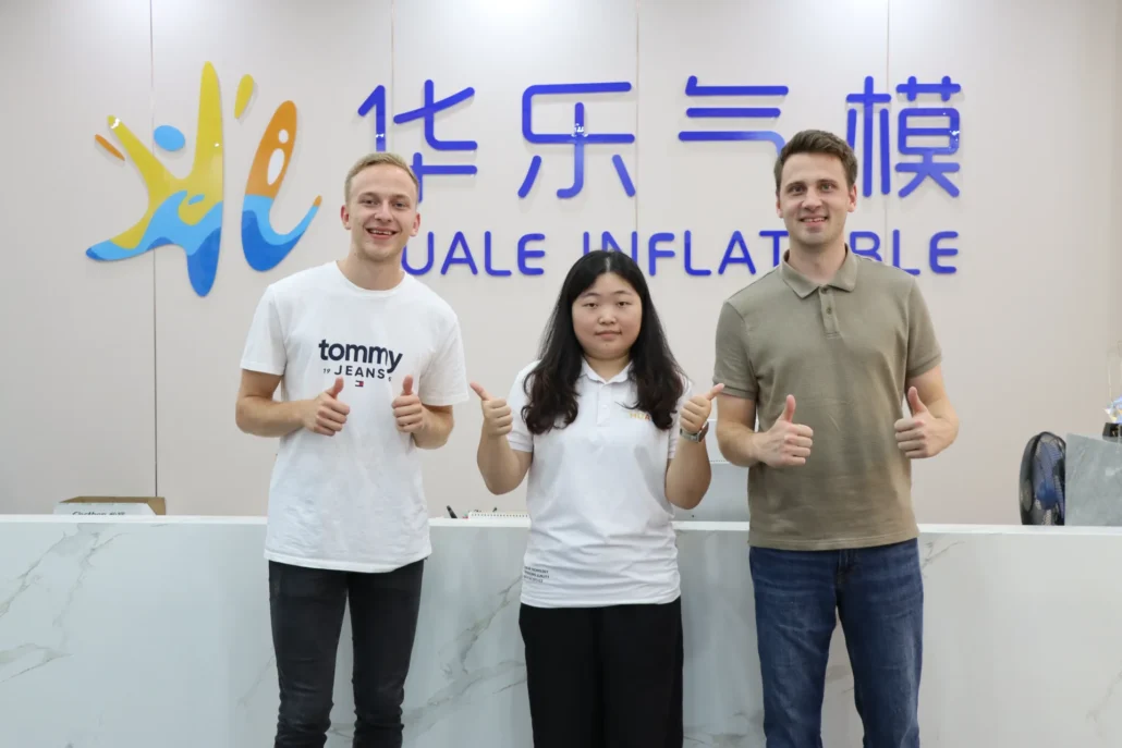 Three individuals posing in front of the Huale Inflatables sign, smiling and giving thumbs up, standing behind a marble reception desk.