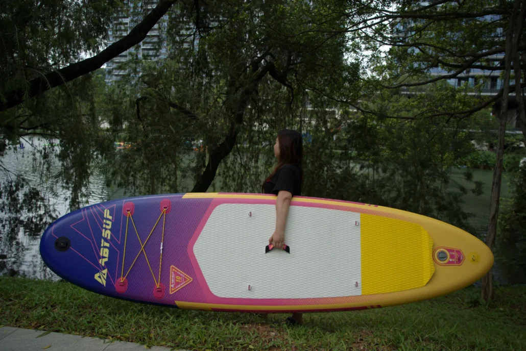 A woman standing near a river holding a vibrant paddleboard with purple, pink, and yellow colors, surrounded by trees.