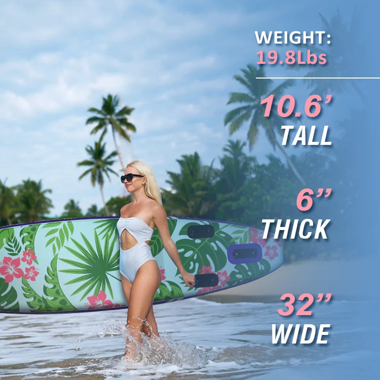 A woman in a white swimsuit carrying a tropical-themed paddleboard with palm trees and flowers at a sunny beach.
