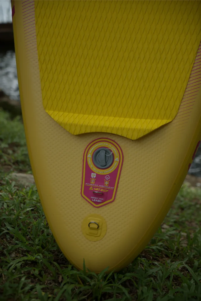 Close-up view of a yellow paddleboard showing its inflation valve, textured surface, and a leash connector resting on grass outdoors.