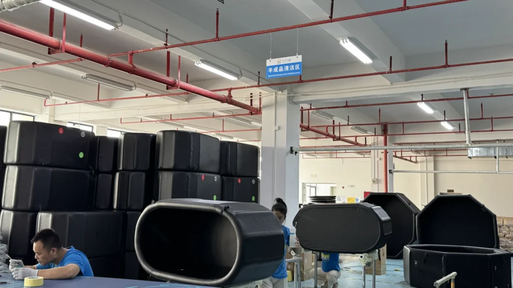 Workers assembling large black storage containers in a factory production line with labeled sections and red pipework overhead.