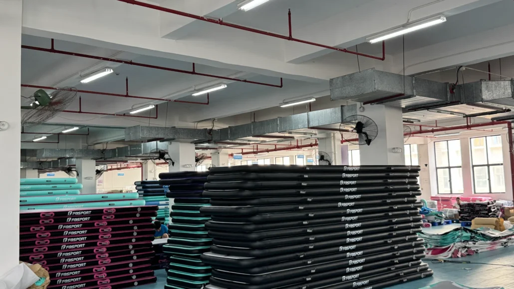 Interior of a factory with stacks of inflatable mats in black and turquoise colors, fans, industrial ventilation, and workers in the background.
