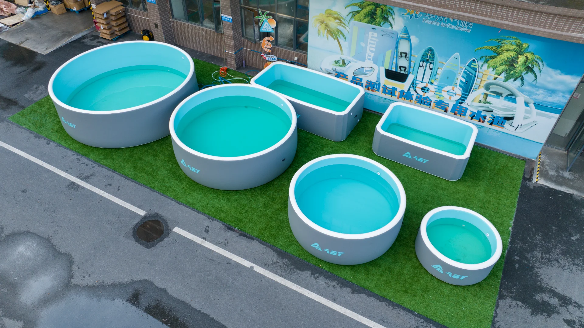 An assortment of large portable swimming pools in various shapes and sizes on green artificial turf with a vibrant backdrop.