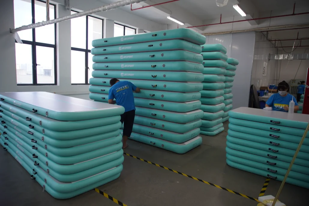 A factory setting with workers assembling stacked turquoise airtrack mats under bright lighting.